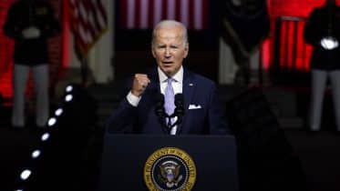 An older man in a dark suit and tie speaks passionately at a podium with the presidential seal, in front of a dimly lit audience with spotlights highlighting red banners. This gathering is of Americans who