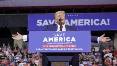 A man speaks at a podium with "Save America" banners at a Trump rally, addressing an audience in an indoor arena. His arms are spread wide as he speaks.