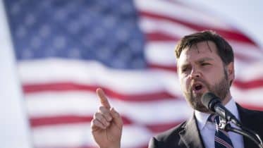 A man delivering a speech, gesturing with his index finger, with a large American flag in the background. He appears focused and earnest, embodying the spirit of an Ohio Republican.