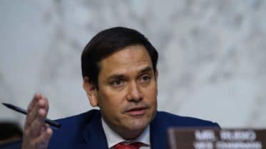 A man in a suit, sitting at a desk with a microphone, looking intently during a discussion. A nameplate reading "Sen. Rubio" is visible in the foreground.