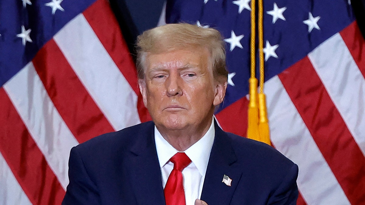President Trump in red tie, dark coat with US flags behind him