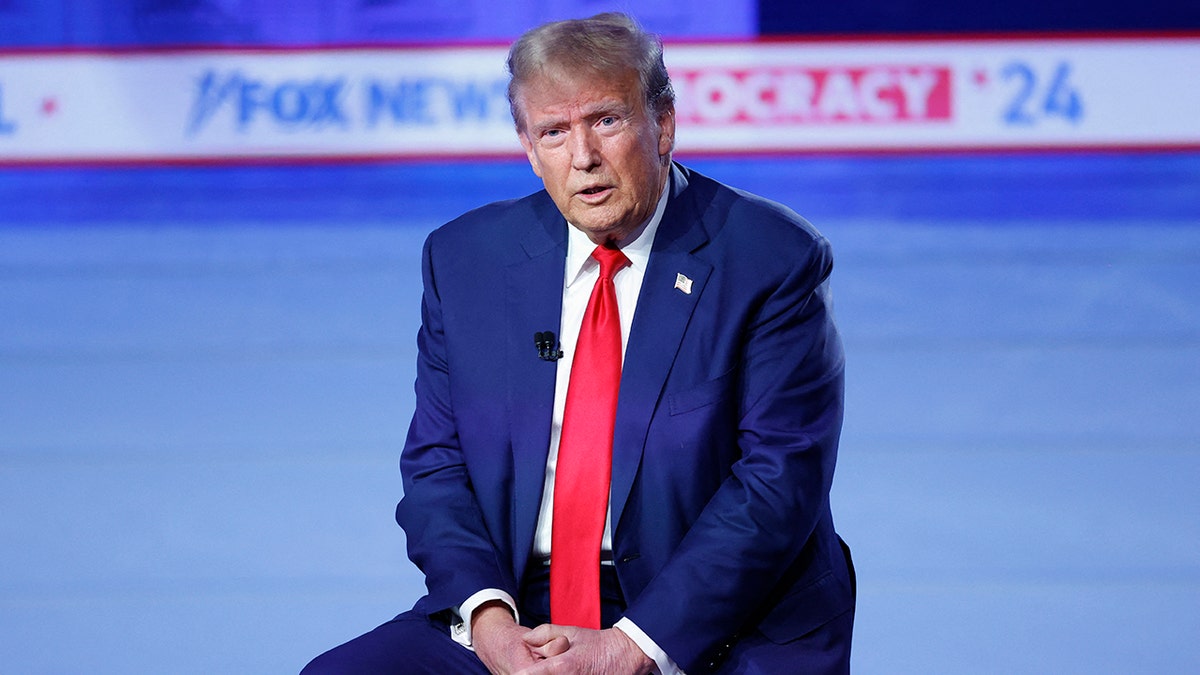 Trump talking, sitting down on stool on stage, folded hands across lap, wearing a navy suit with a bright red tie