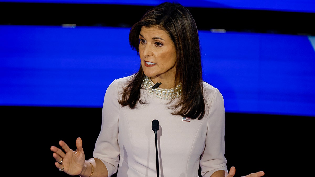 Nikki Haley wearing a light pink dress, with pearl necklace speaking on stage with hands open