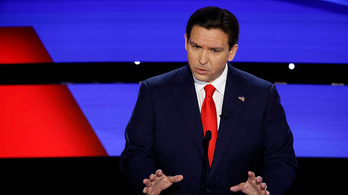 Ron DeSantis wearing navy suit, bright red tie, arms out and hands open, mid sentence