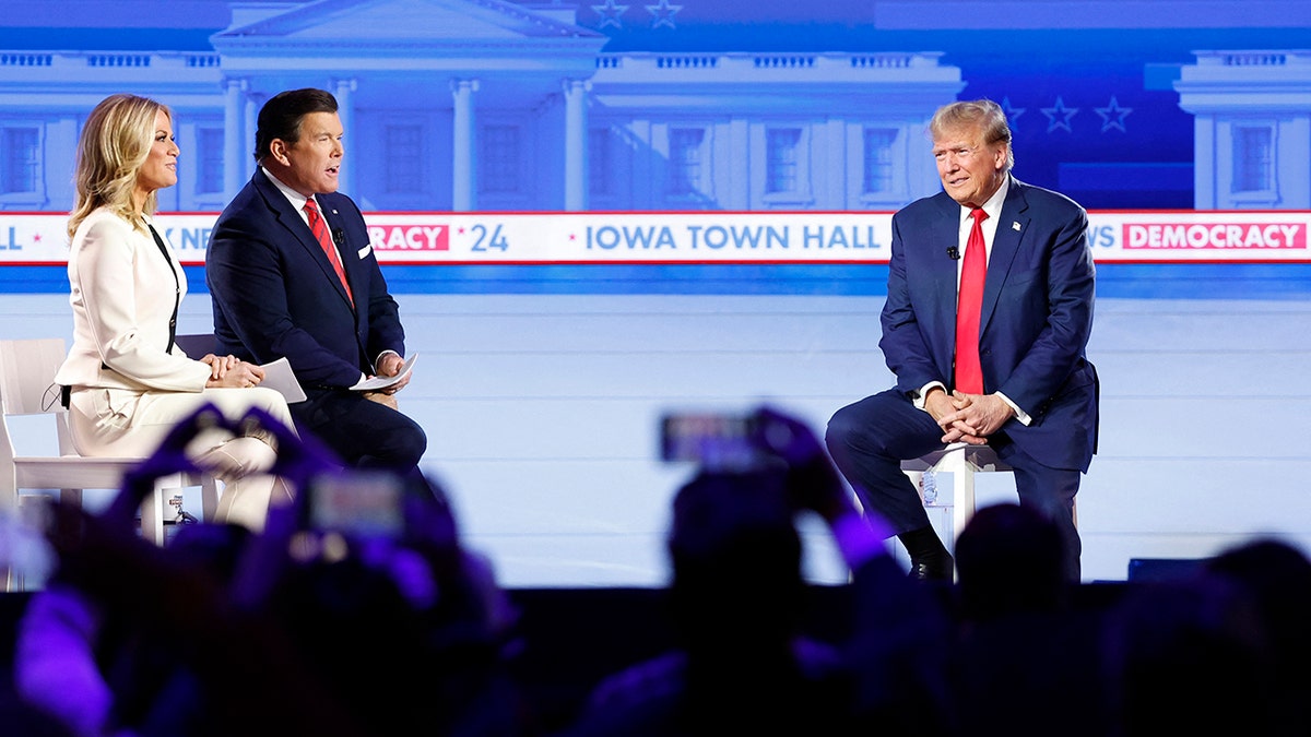 Fox News hosts Martha MacCallum and Bret Baier talking with Trump on stage during Fox News town hall, audience holding up heart sign with hands and phone to record