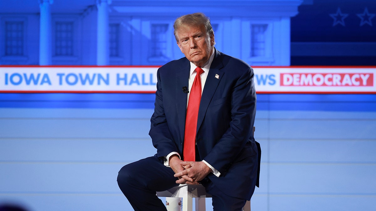Trump with serious look on his face, sitting down on stool on stage, folded hands across lap, wearing a navy suit with a bright red tie