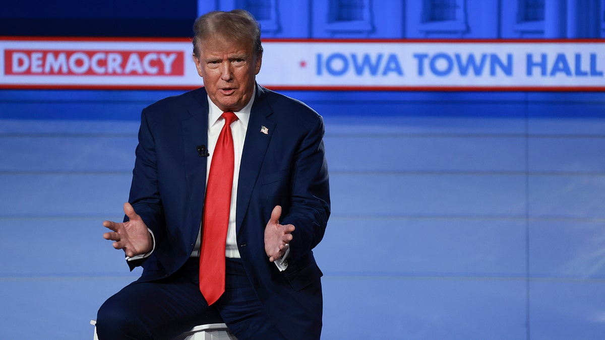 Trump mid sentence, hands open, wearing a navy suit with a bright red tie, sitting on stool on stage, Democracy, Iowa Town Hall banner in background