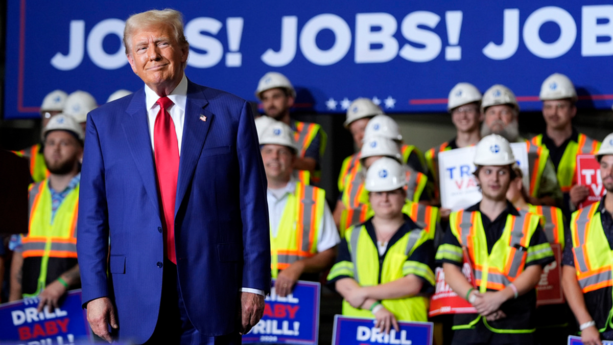 Donald Trump in blue coat, red tie, "Jobs!" sign behind him