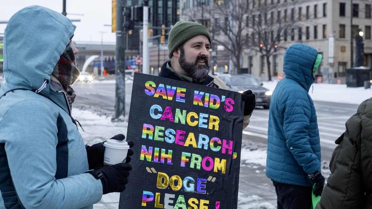 The "No Kings on Presidents Day" protest is pictured on Feb. 17, 2025, in Detroit in response to what they say are President Donald Trump and Elon Musk's undemocratic actions. Elliot Stephens carries a sign about cancer research. His teenage daughter has brain cancer and is currently part of a clinical trial.