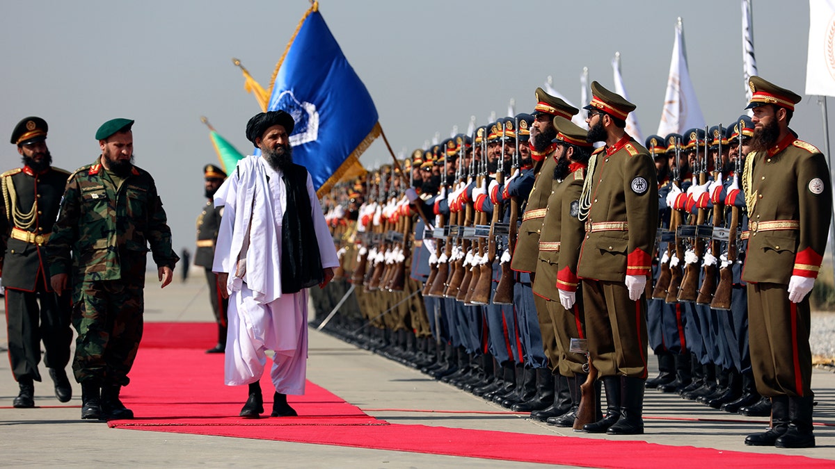 A senior Taliban official inspects a line of uniformed soldiers standing at attention during a military parade in Afghanistan. The official walks on a red carpet flanked by armed guards, with ceremonial troops holding rifles in formation. A large blue flag and other national or organizational banners are visible in the background.