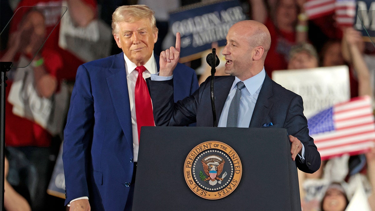 Stephen Miller speaks at a podium with President Donald Trump standing behind him and smirking