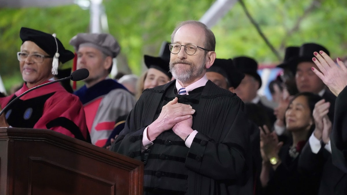 Harvard President Alan Garber places his hands over his heart while standing at the podium during the university’s commencement ceremony, as faculty in academic regalia applaud behind him.
