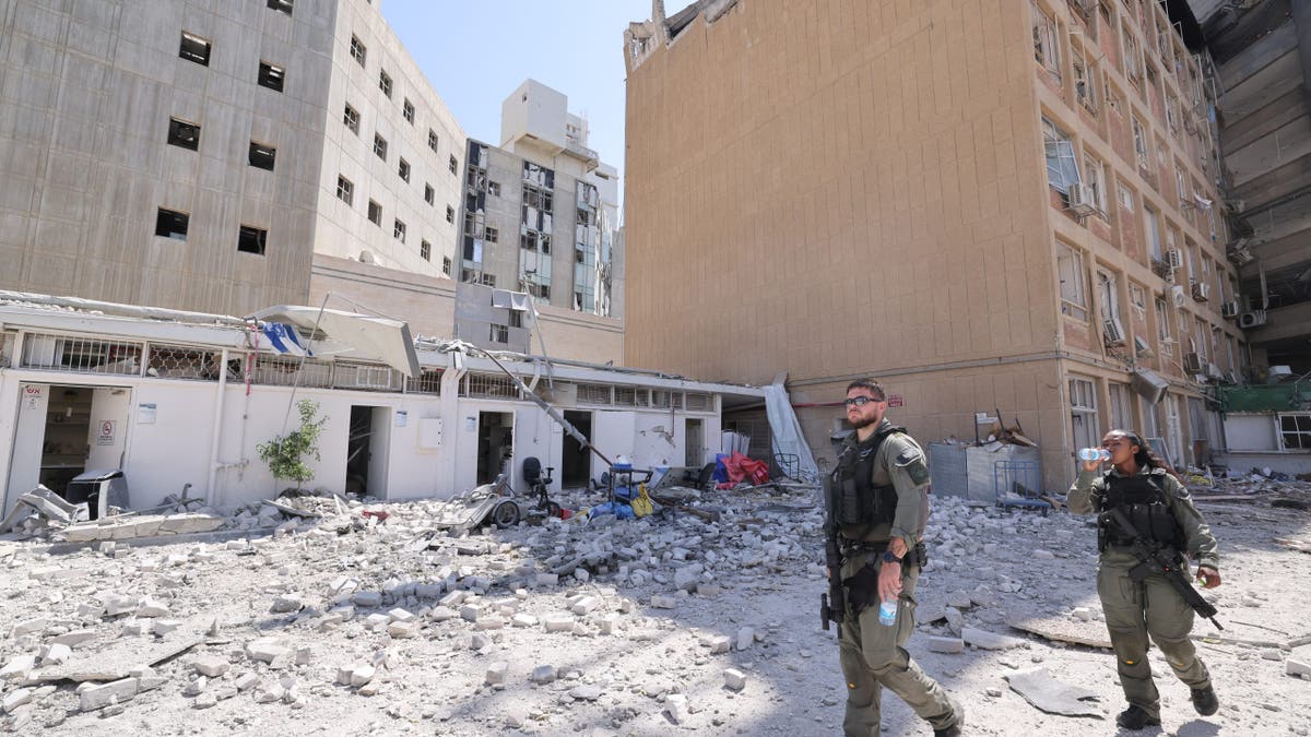 Two armed Israeli security personnel patrol a debris-strewn area outside a damaged building hit during conflict in southern Israel.