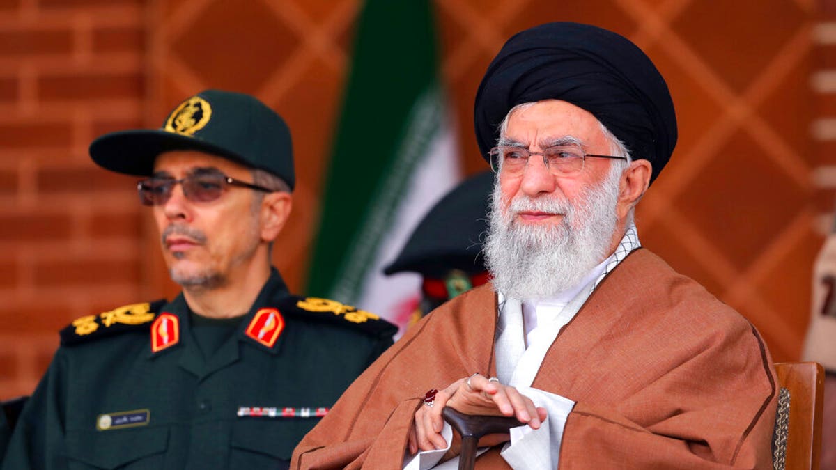 Supreme Leader Ayatollah Ali Khamenei sits in a chair and looks on while an Iranian solider stands behind him