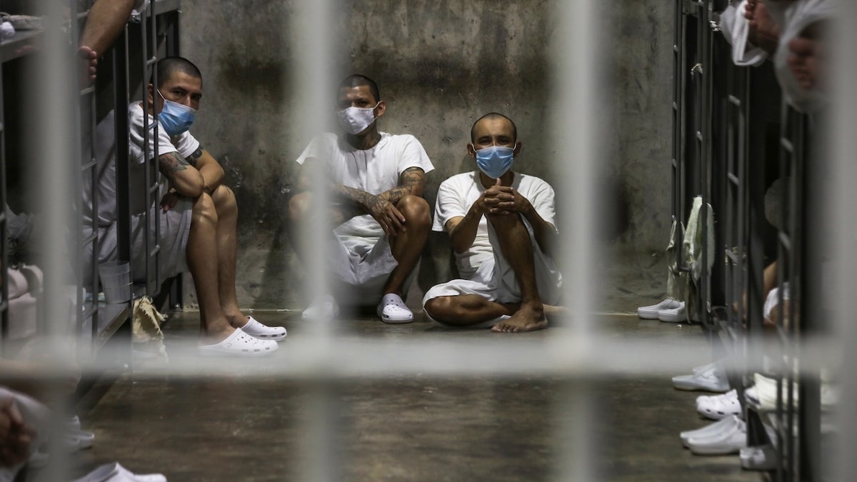 Detainees in white clothing and surgical masks sit inside a crowded Salvadoran prison, photographed through metal bars.