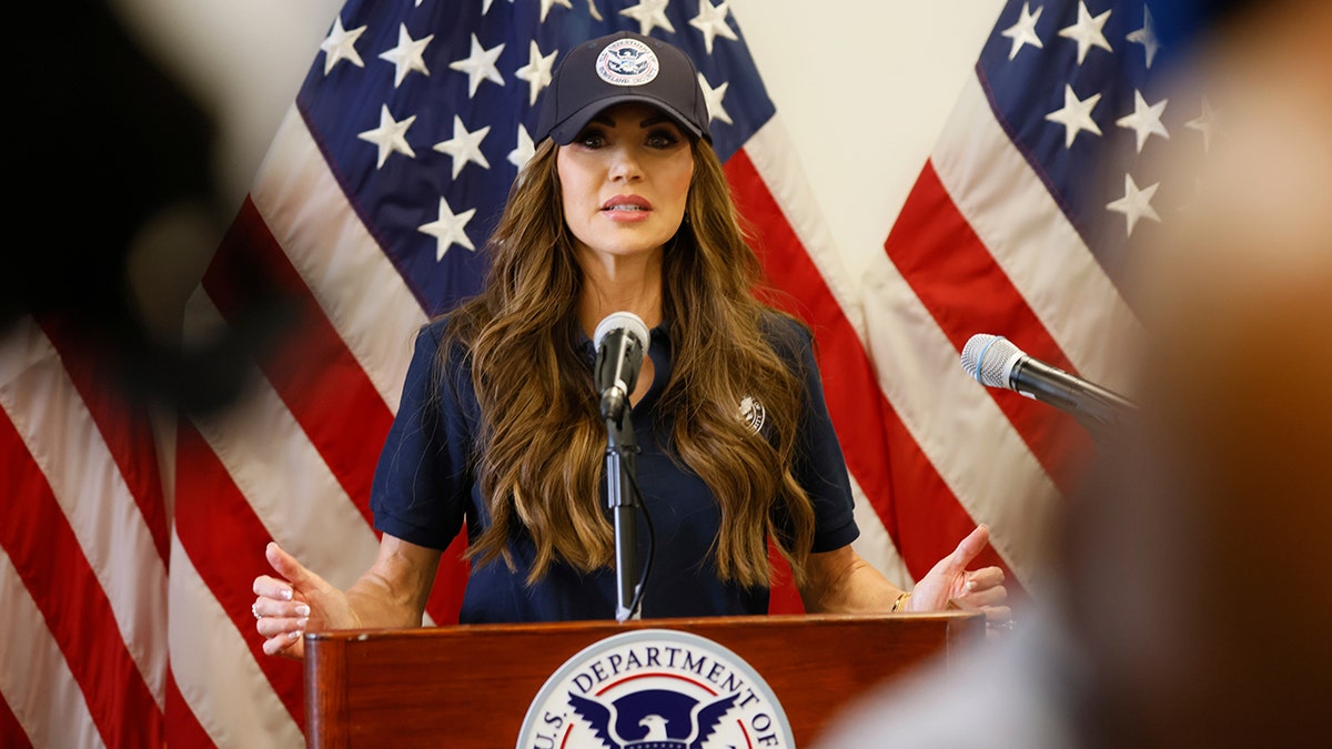 Homeland Security Secretary Kristi Noem speaking at a podium wearing a blue DHS hat
