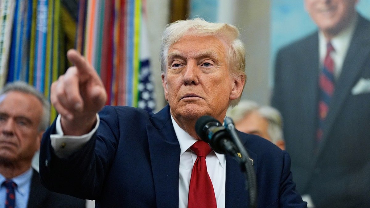 President Donald Trump wearing blue suit and a red tie while pointing at a reporter in the Oval Office.