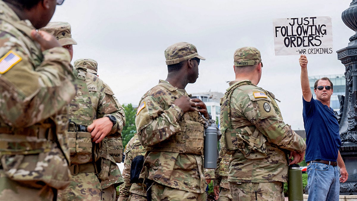 Soldiers look as protester holds sign reading "Just following orders - War criminals"