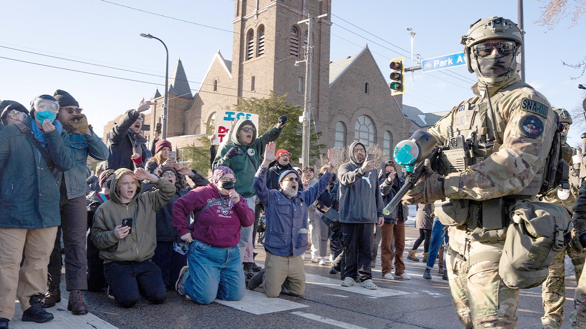 Community members, some kneeling with hands raised, shout at federal agents on a Minneapolis street amid heightened tensions.