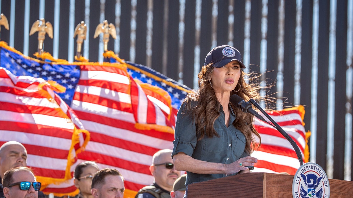Kristi Noem during immigration presser in Arizona speaking at a microphone with American flags in the background