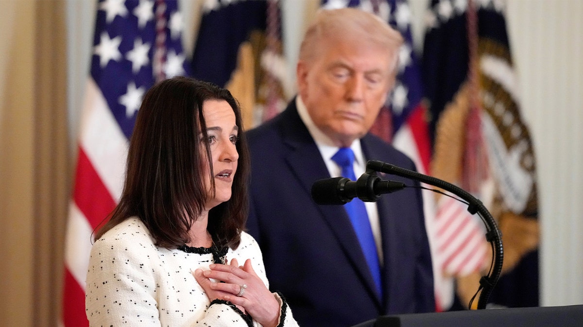 A woman speaks at a podium in the East Room of the White House as President Donald Trump looks on.