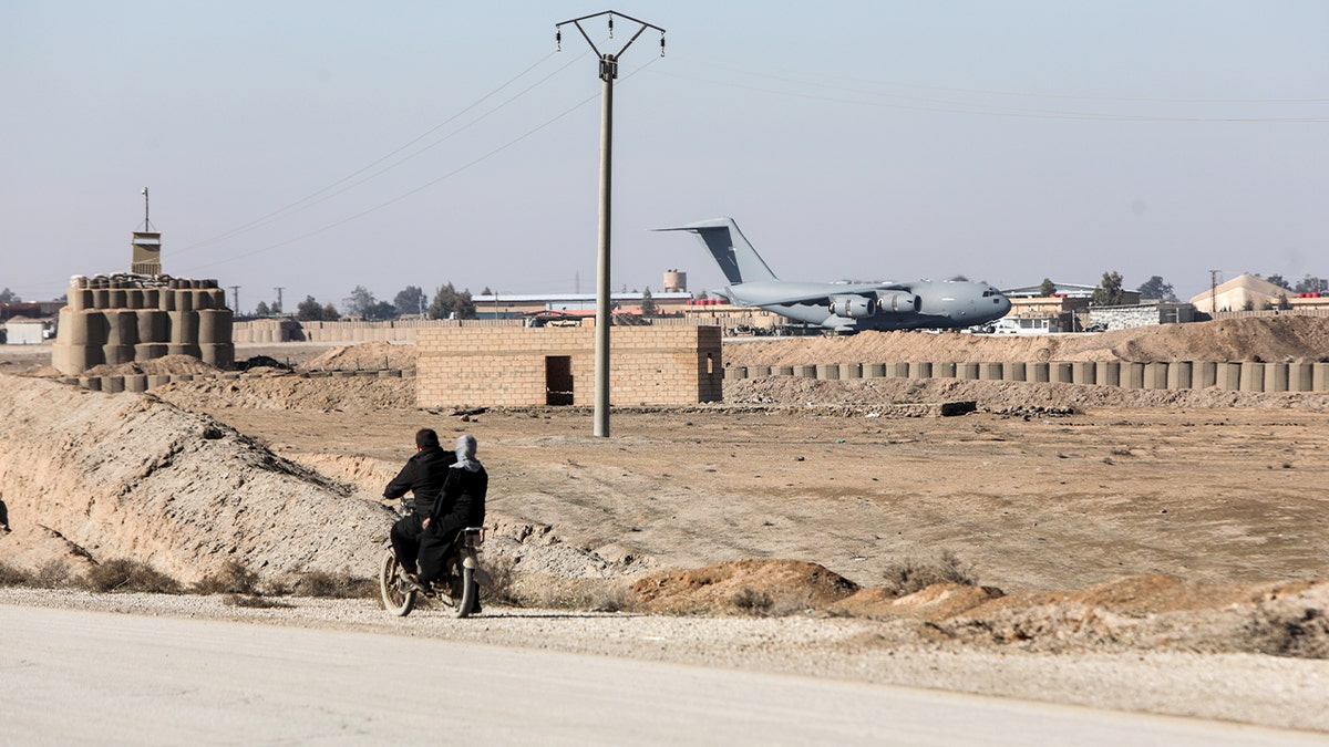 Two people ride a motorbike along a road near a military base where a coalition aircraft is visible overhead in Al-Shaddadi.