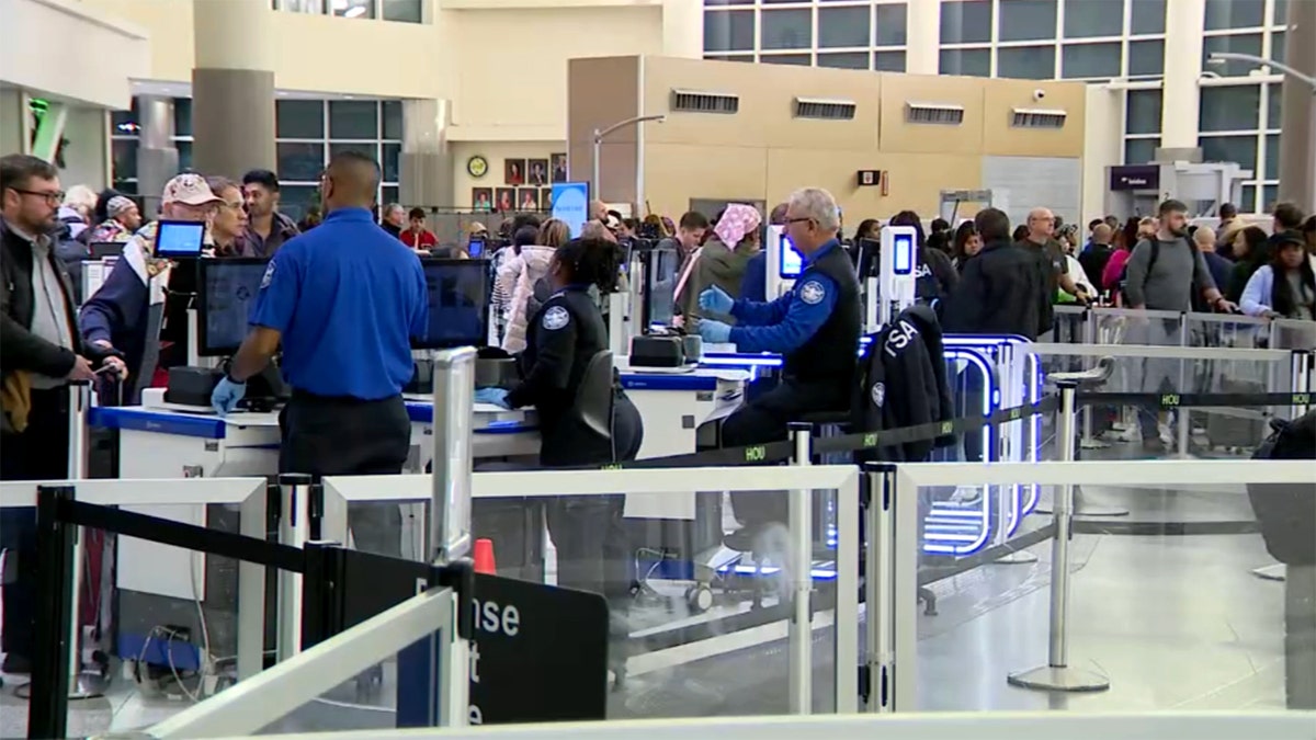 TSA agents screening passengers at airport security checkpoint with travelers in line and luggage scanners in a busy terminal.