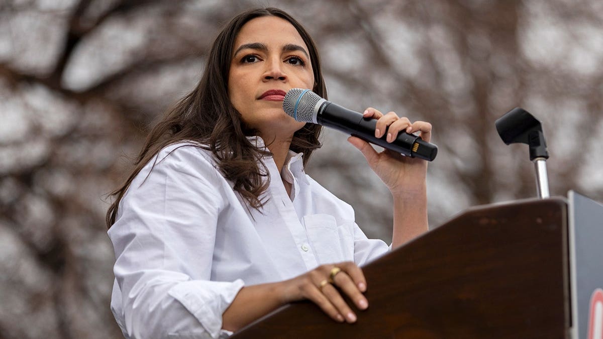 Rep. Alexandria Ocasio-Cortez holding a microphone while standing at a podium