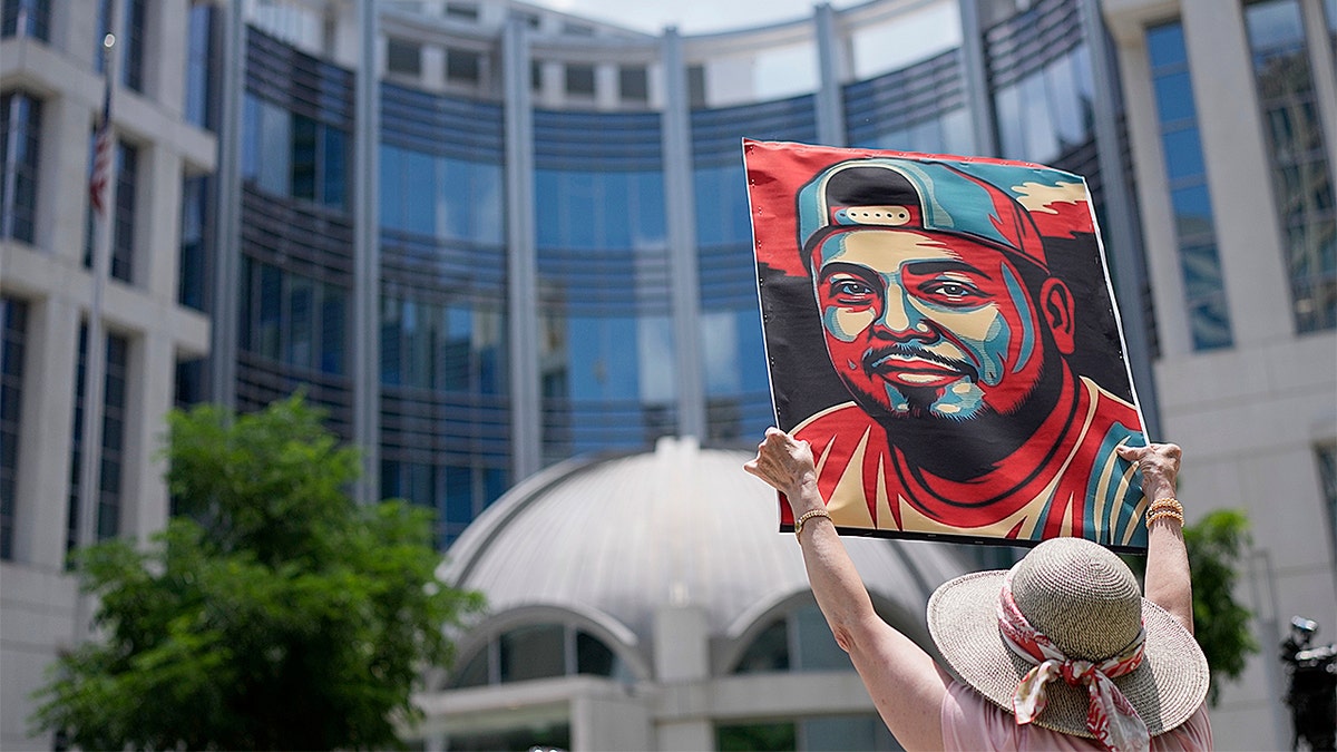 Woman holds up portrait of Kilmar Abrego Garcia outside a building.