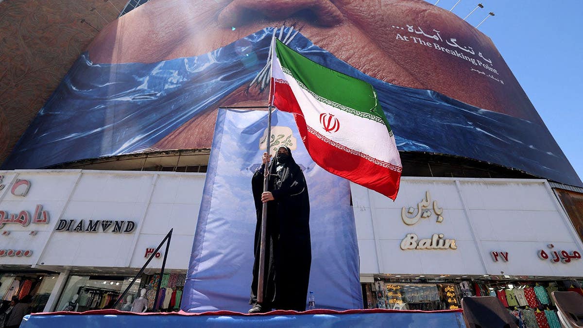 A woman holding the Iranian flag standing in front of an anti-US billboard in Tehran