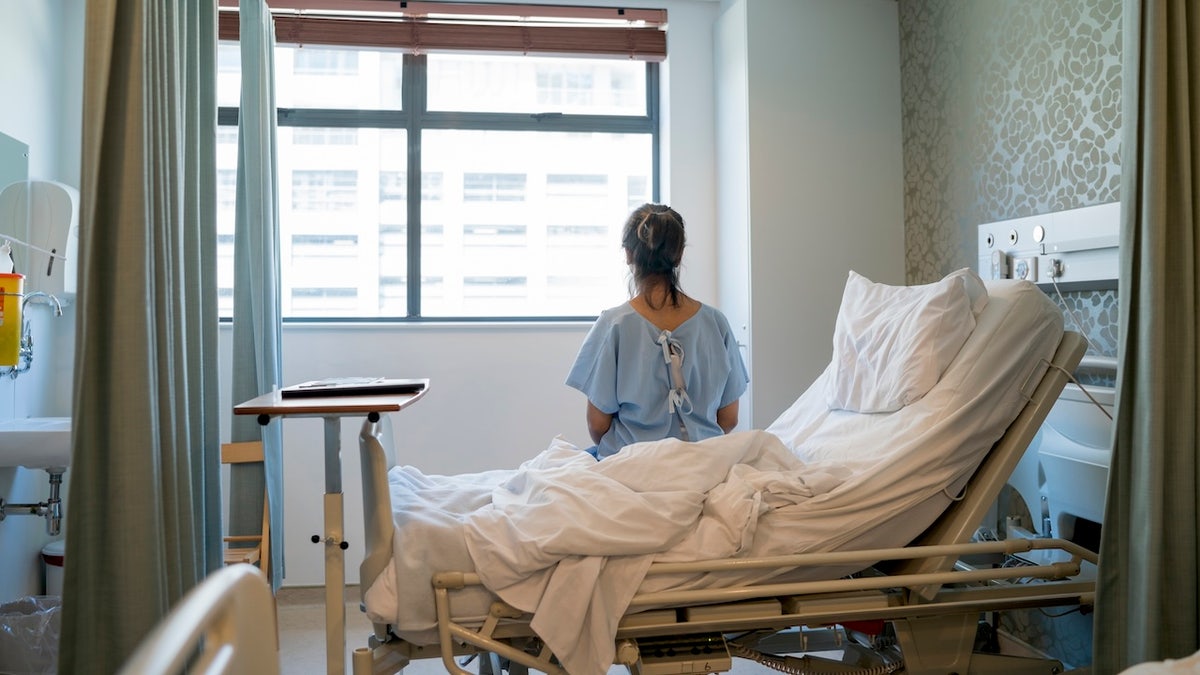 Female patient sitting on hospital bed wearing hospital gown in ward