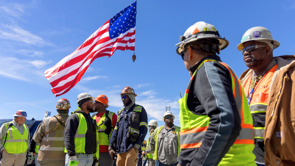 Construction workers work on a site while an American flag waves overhead.