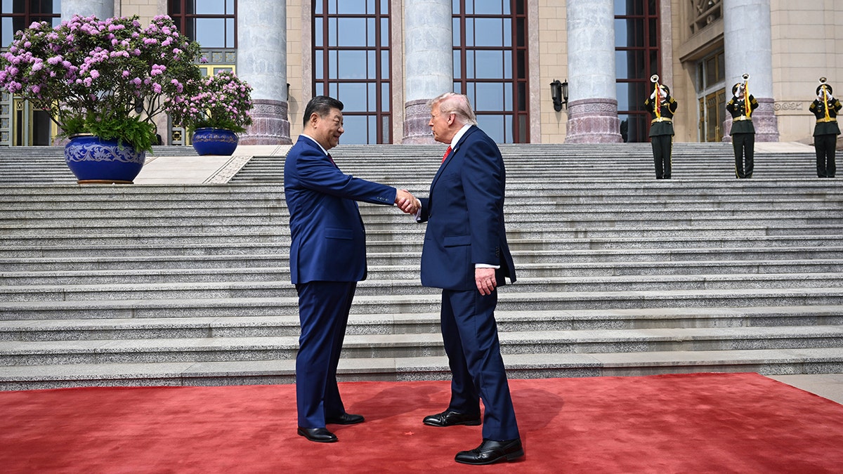 Xi Jinping and Donald Trump shake hands while standing on a red carpet in front of large stone steps.