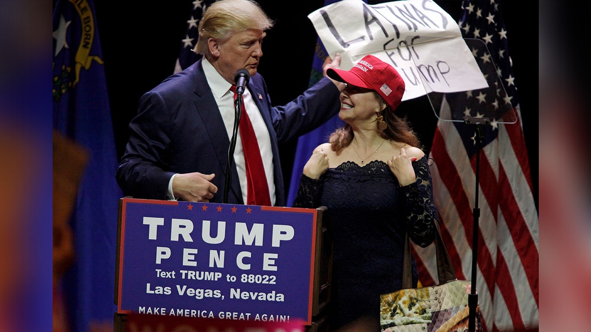 Woman on stage with Donald Trump carrying a "Latinos for Trump" sign.
